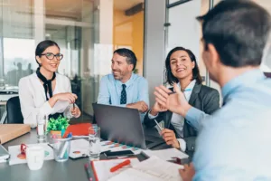 Four business colleagues smiling during a conversation; one man gestures while speaking, while others listen and smile.