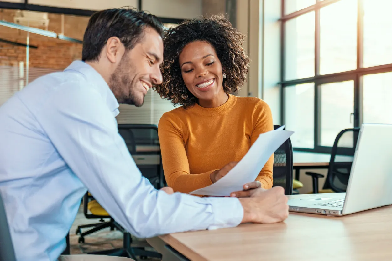 A man and a woman sitting at a desk in an office, reviewing documents together.