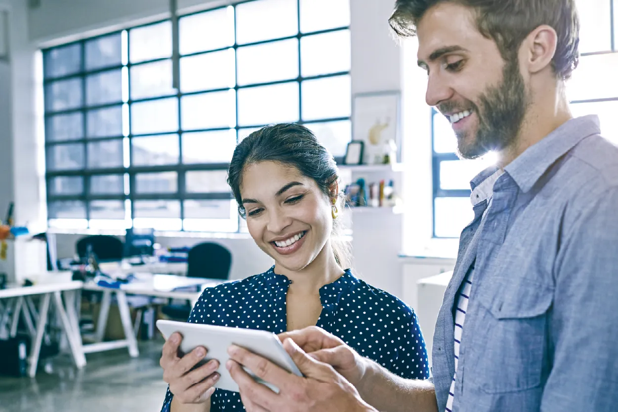 Smiling male and female coworkers standing next to each other, holding one tablet, with the man pointing at the screen.