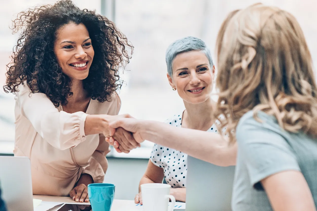 Two women shaking hands across the table in a business meeting, while another woman smiles at them from the opposite side.