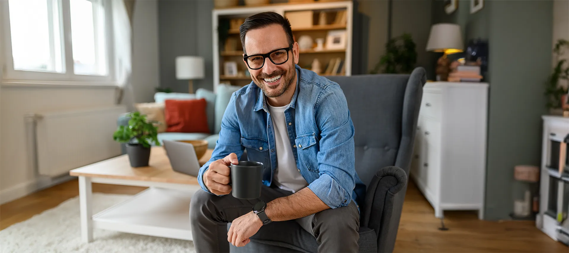 Smiling man seated in a chair in his living room. He's holding a coffee cup, looking at the camera.