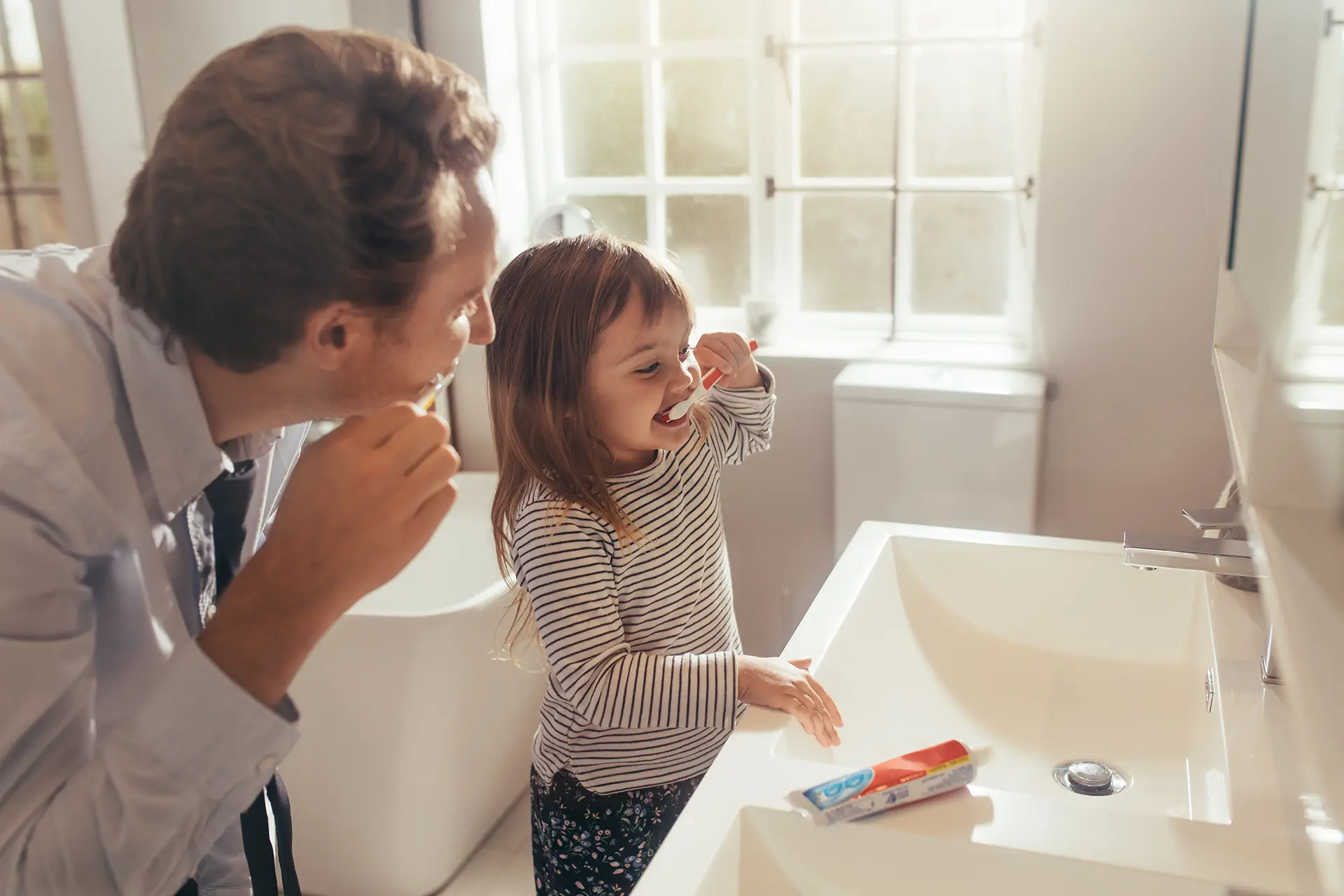 Young daughter and dad brushing their teeth together at the bathroom sink.