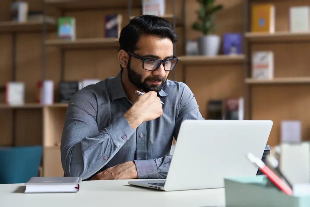 Man wearing glasses seated at his desk looking at his laptop. He has his hand under his chin.