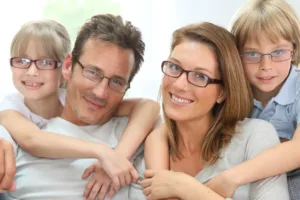 Family of four on a couch, all wearing glasses. Daughter hugs dad’s neck; son hugs mom’s neck.