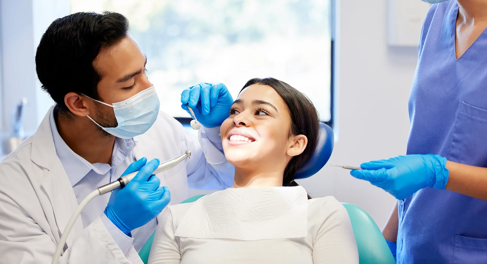 A female patient with a bright smile in a dental chair, with a male dentist and a female assistant examining her teeth.