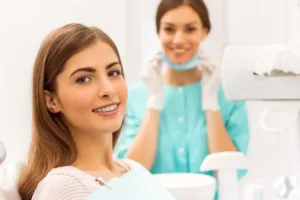 Teenage girl with braces sitting in an orthodontist chair, looking at the camera. Orthodontist stands in the background.
