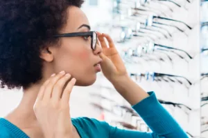 Woman trying on glasses in an optical store, standing in front of a display wall and looking at herself in the mirror.