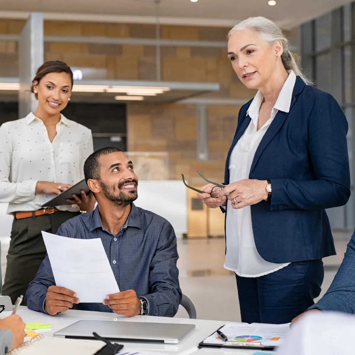 A business meeting in an office with men and women. A woman stands holding her glasses, listening, as others look at her.