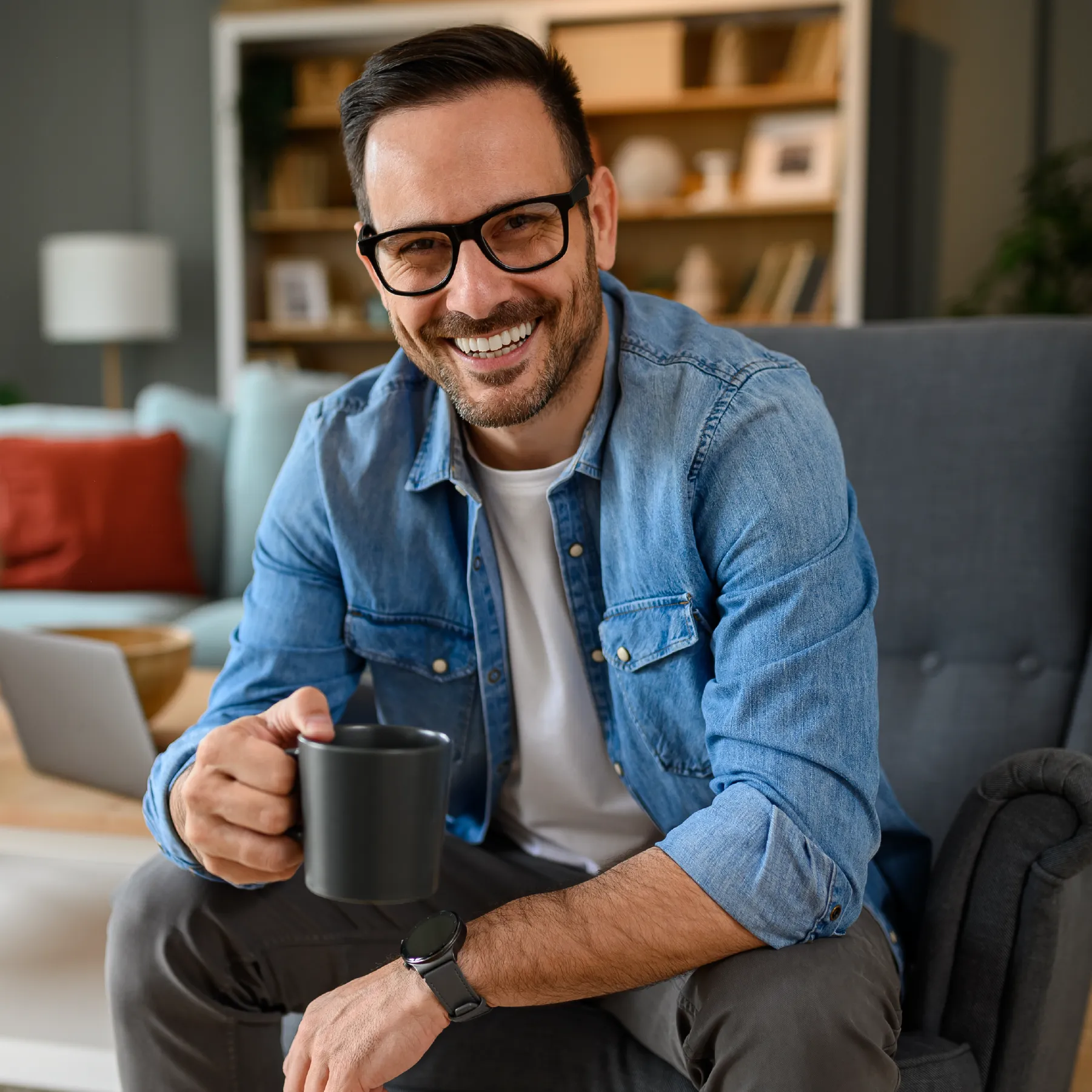 Smiling man seated in a chair in his living room. He's holding a coffee cup, looking at the camera.