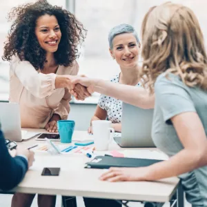 Two women shaking hands across the table in a business meeting, while another woman smiles at them from the opposite side.