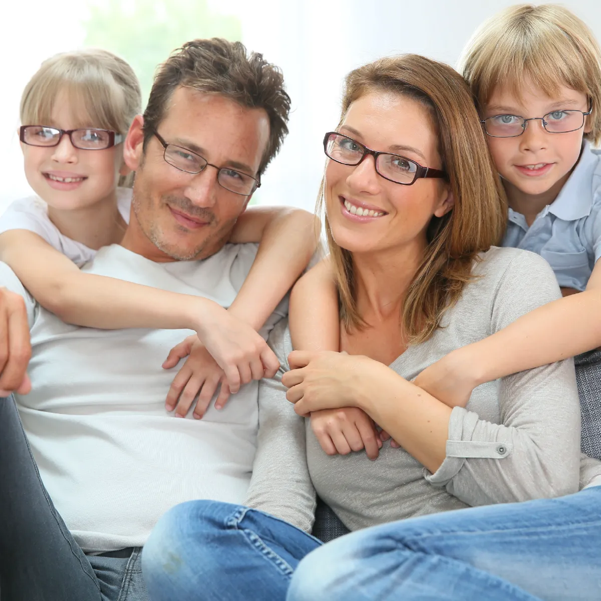 Family of four on a couch, all wearing glasses. Daughter hugs dad’s neck; son hugs mom’s neck.