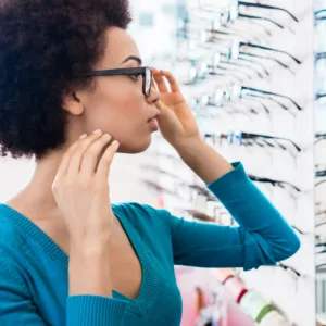 Woman trying on glasses in an optical store, standing in front of a display wall and looking at herself in the mirror.