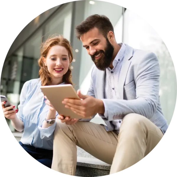 Smiling man and woman sitting outdoors, looking at a tablet together while holding a smartphone and chatting.