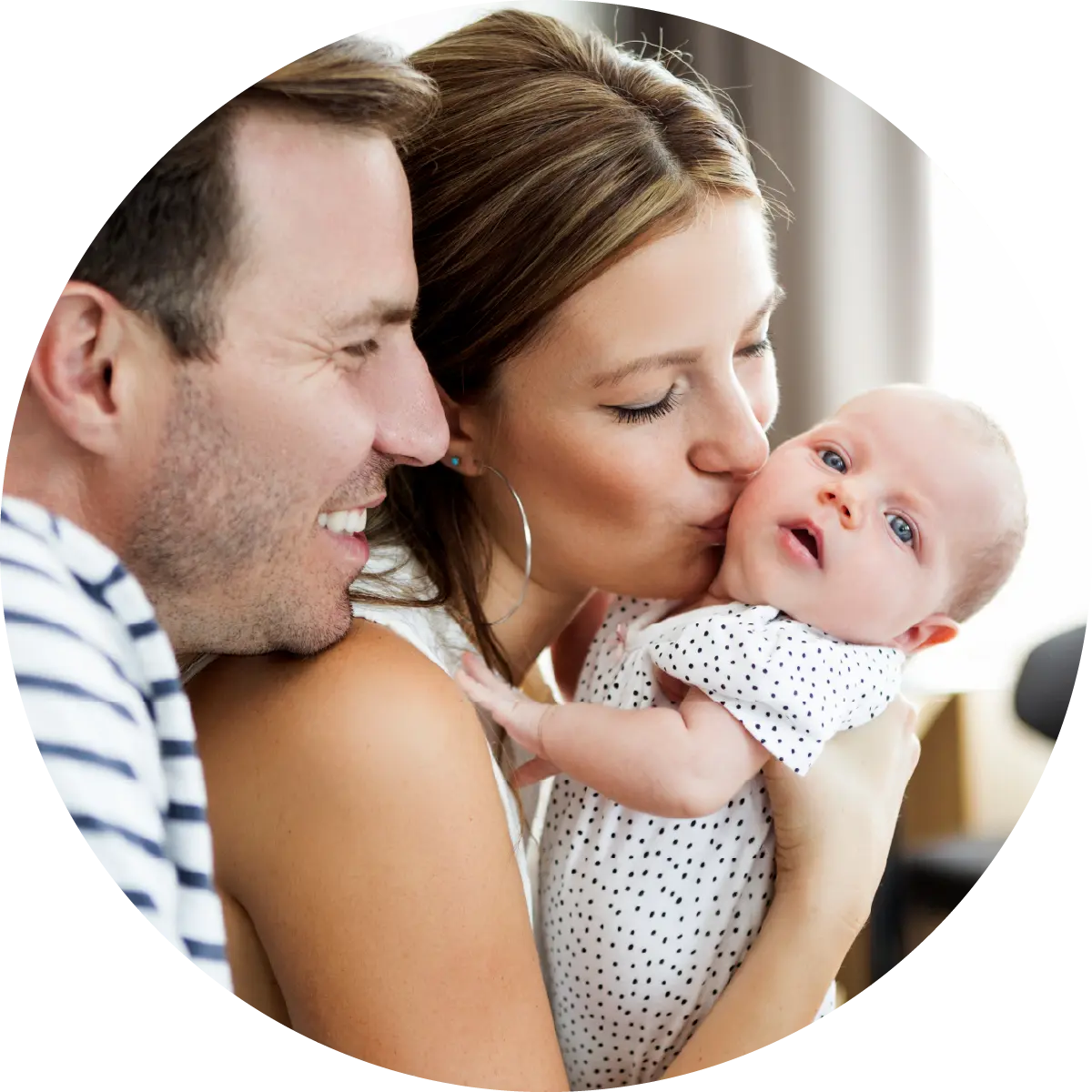 A mother kisses her baby while the father watches with a warm smile and affectionate gaze.