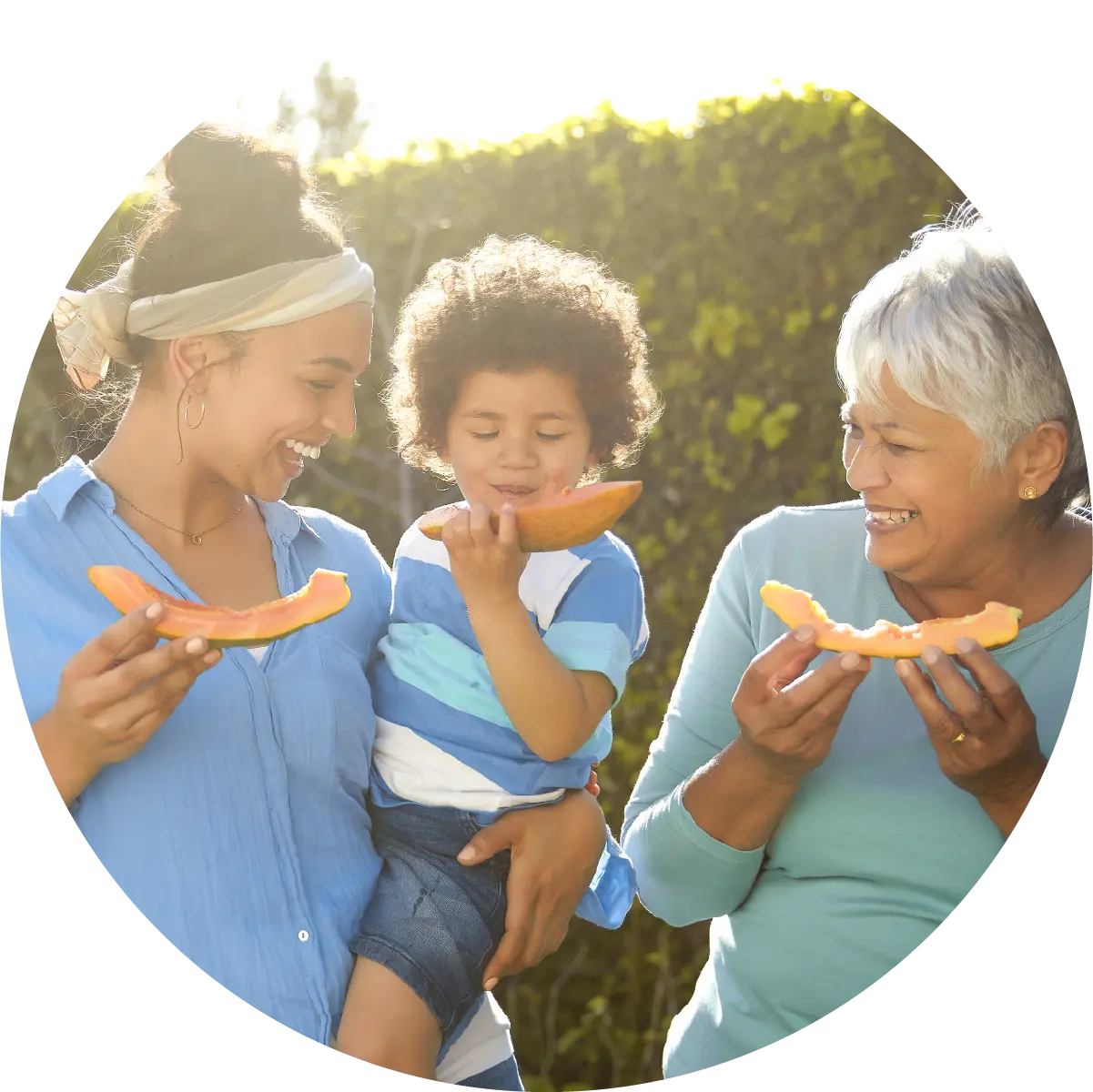 Grandmother, mother, and young son outside, eating cantaloupe wedges with their hands.