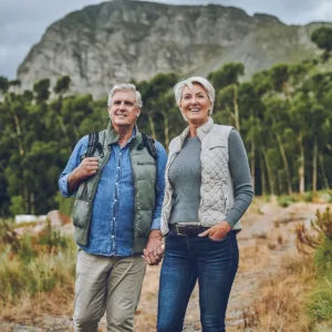 Senior couple standing outdoors holding hands with mountains in the background, dressed for hiking.