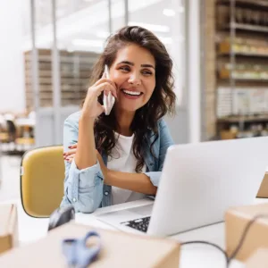 A woman sitting at a desk in a warehouse. She is talking on her cell phone with an open laptop in front of her.