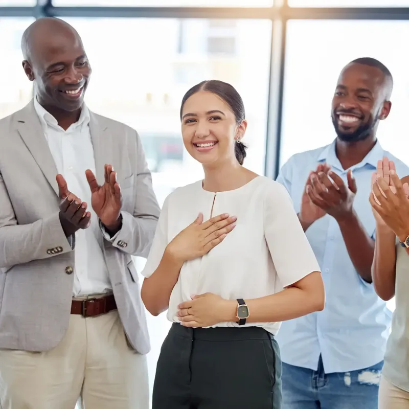 Employees celebrating a woman who stands in the center, smiling with her hand on her heart. Others are clapping around her.