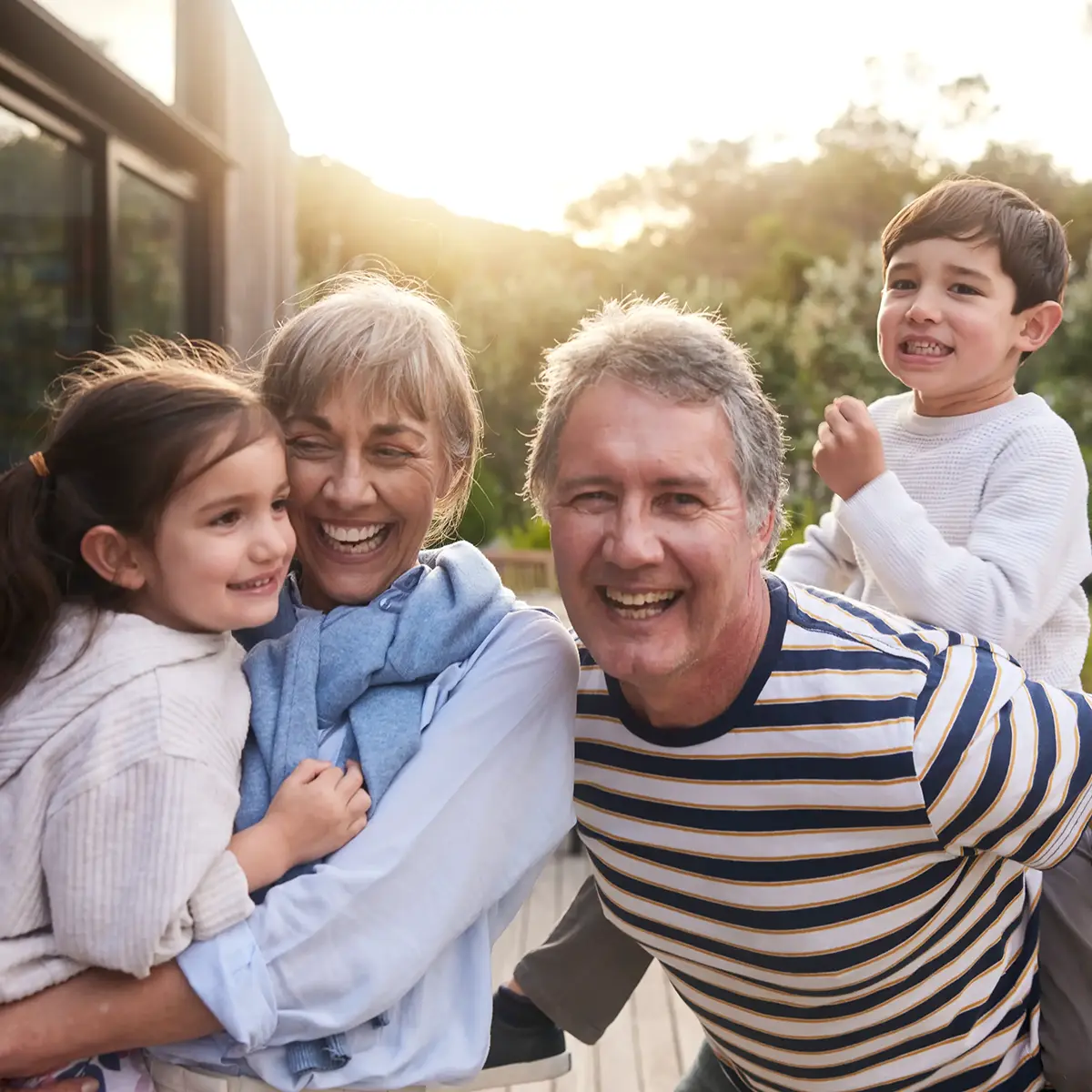 Grandma holding her granddaughter and grandpa giving his grandson a piggyback ride, all smiling outdoors.