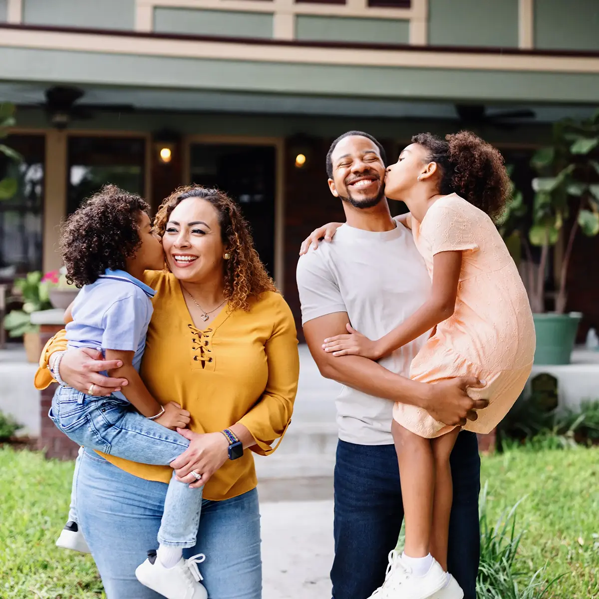 Mom and dad standing outside, each holding a child who is kissing their cheek.