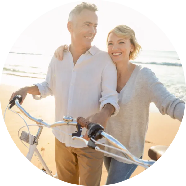 Smiling middle-aged couple walking with bicycles along a beach, enjoying a sunny day together by the ocean.