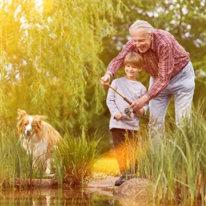 Elderly man teaches a child how to fish at a pond, with a dog nearby and greenery in the background on a sunny day.