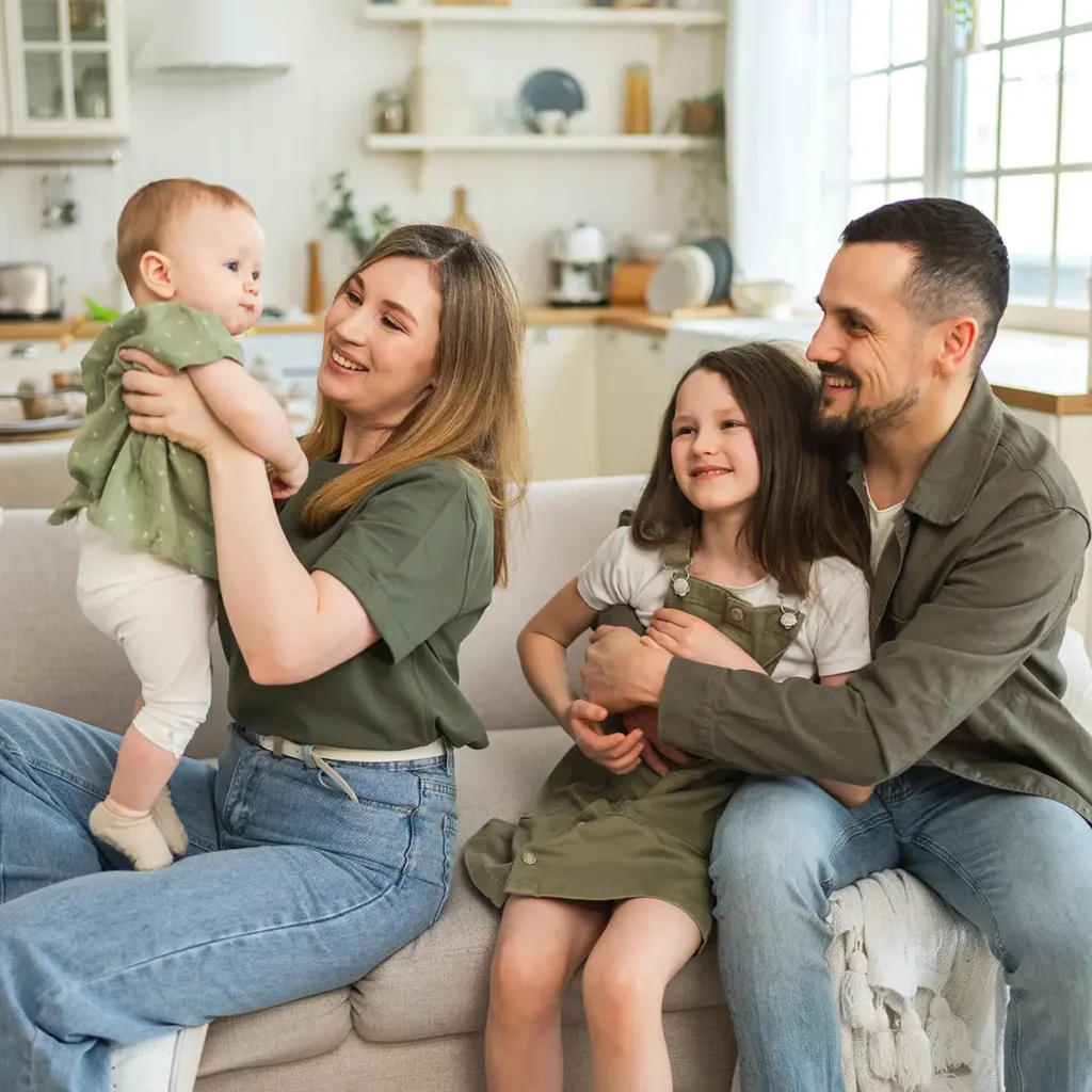 A young family sitting on a couch. Dad has his arms around his daughter, while mom holds a baby on her lap.