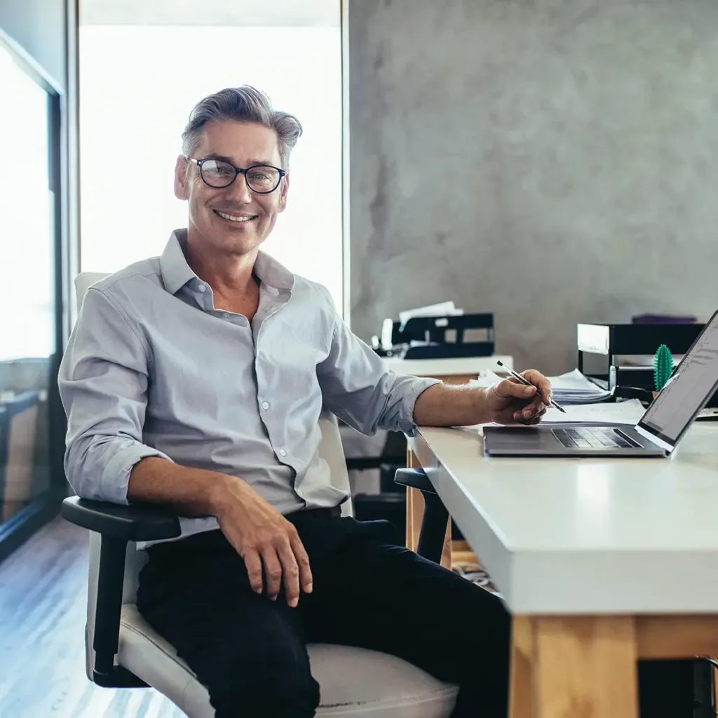 Smiling man wearing glasses is seated at his desk and working on a laptop in his office.