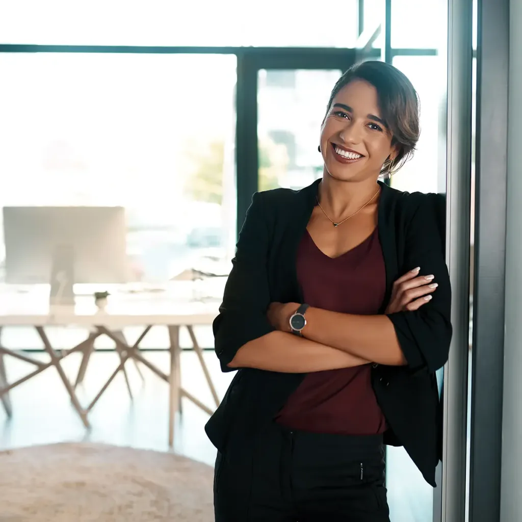 A professional woman stands in her office doorway, smiling with her arms folded across her chest.