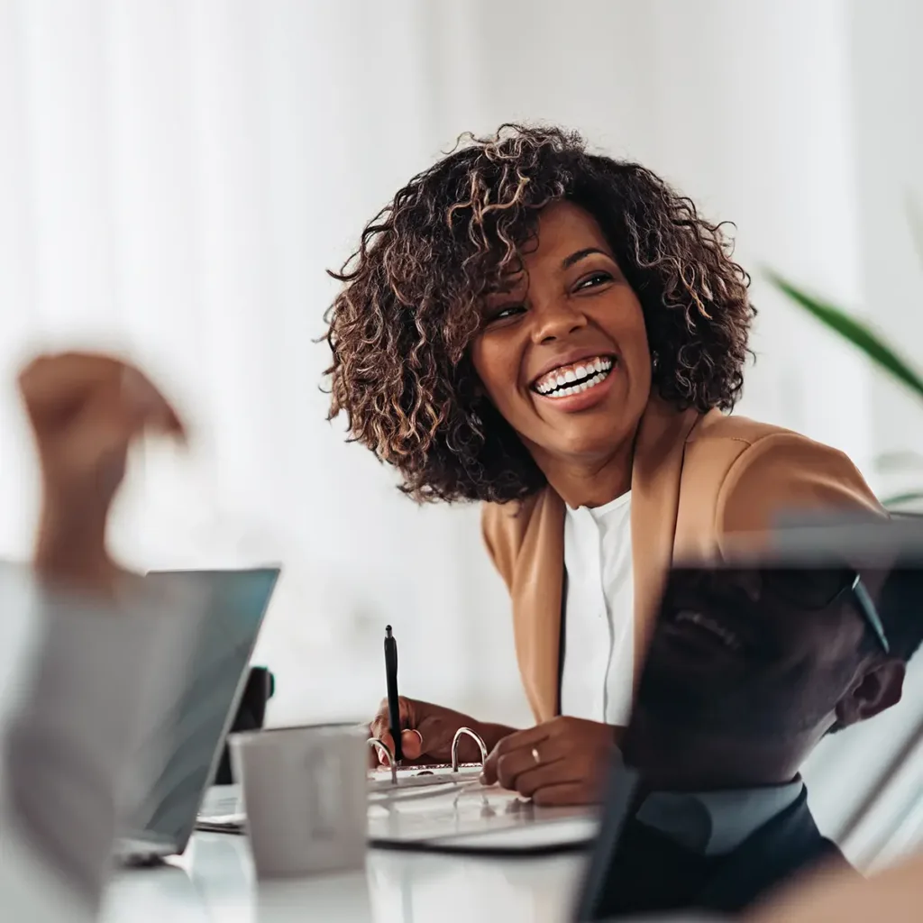 Smiling woman sitting at a conference table with notebook and pen. She is looking to her right at someone who is out of frame.