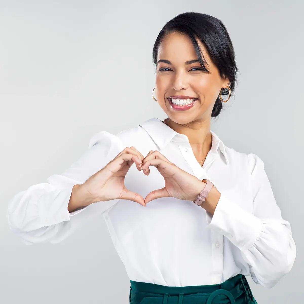 A smiling woman in a white shirt forms a heart shape with her hands.