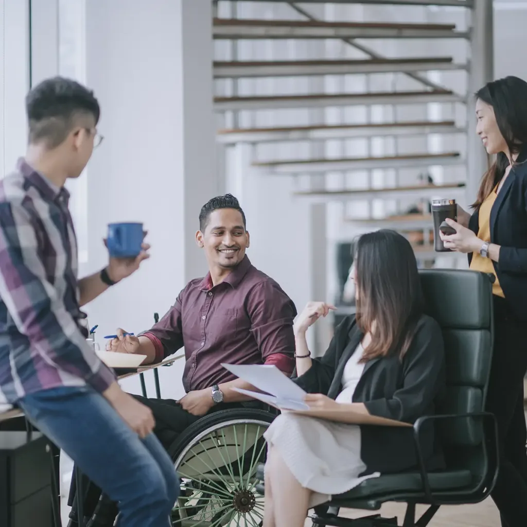 Four coworkers at the office, including a man in a wheelchair and a woman seated in an office chair, standing and smiling together.