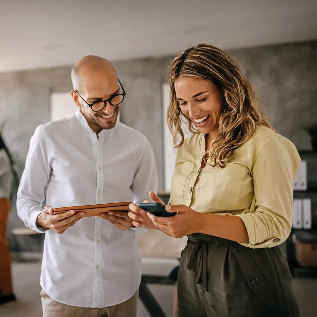 Smiling man and woman standing together; she is holding a phone, he is holding a tablet, and both are looking at her phone.