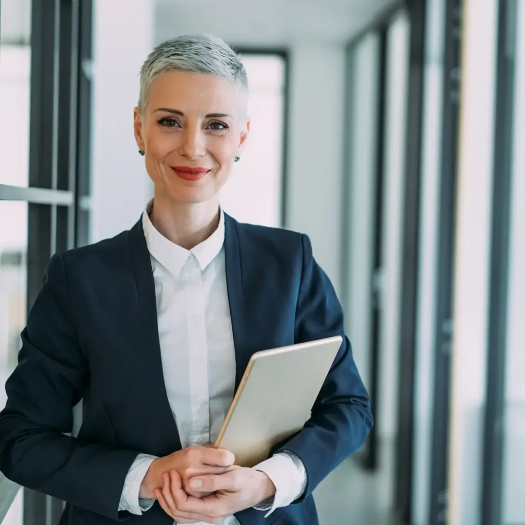 Professional businesswoman stands in an office hallway, smiling and holding a laptop in her arms.