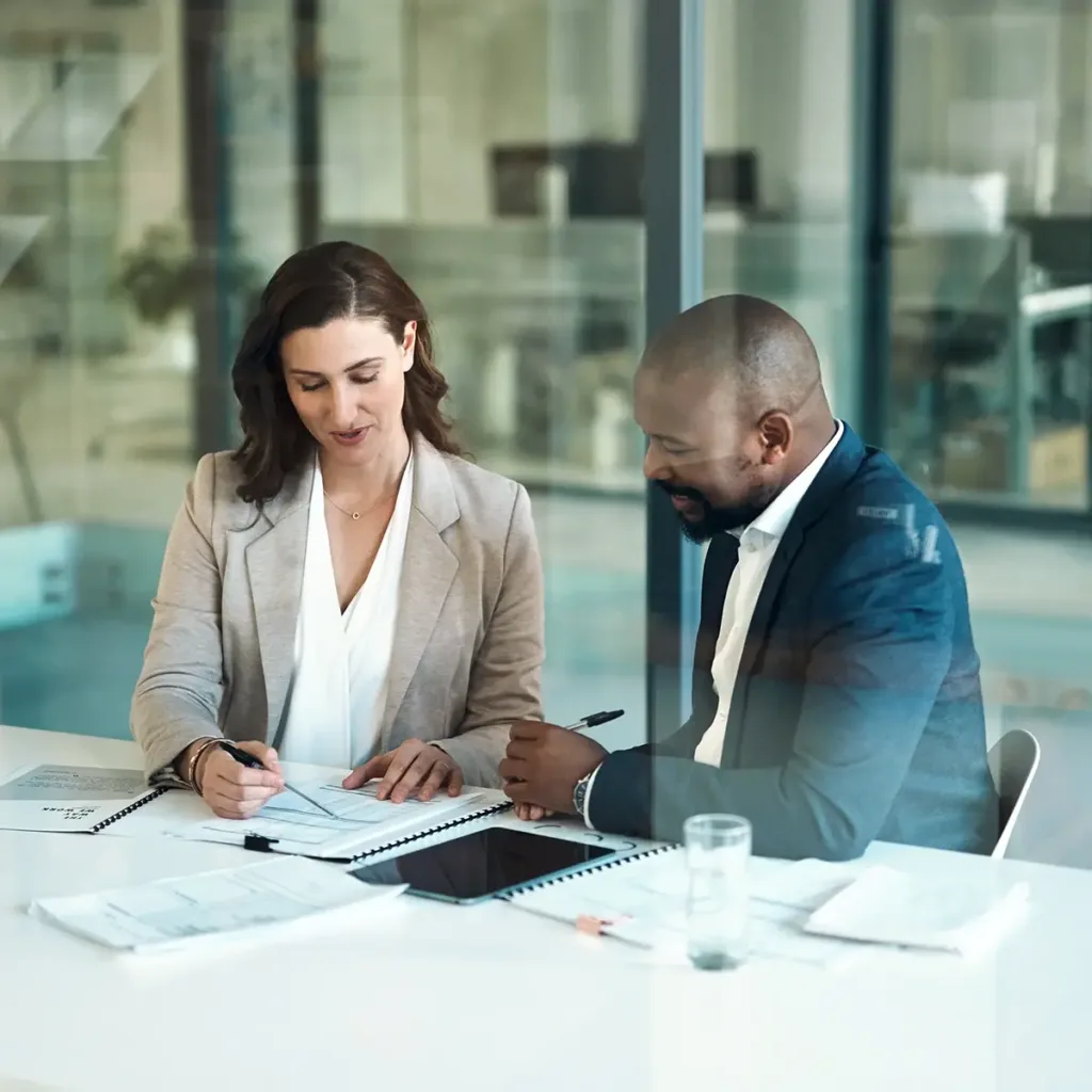 Man and woman in business suits sitting side by side at a table, reviewing paperwork together.