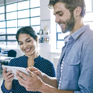 Smiling male and female coworkers standing next to each other, holding one tablet, with the man pointing at the screen.