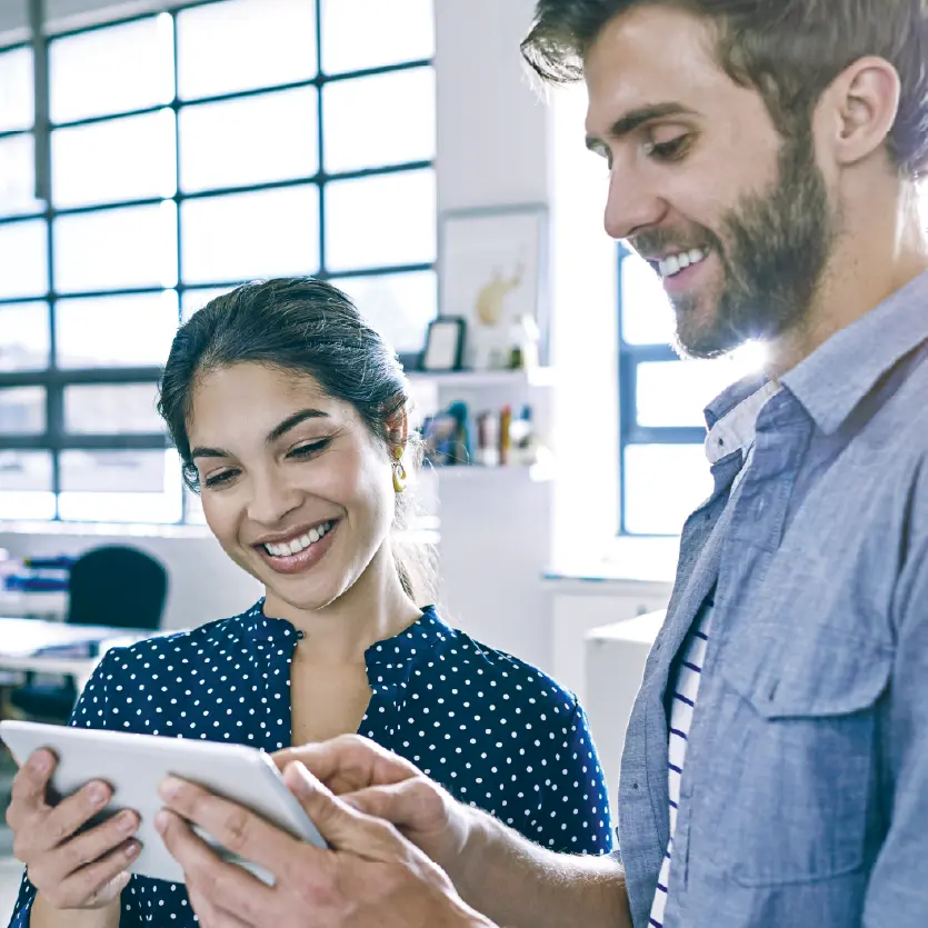 Two smiling colleagues are looking at and interacting with a tablet device in a bright office setting.