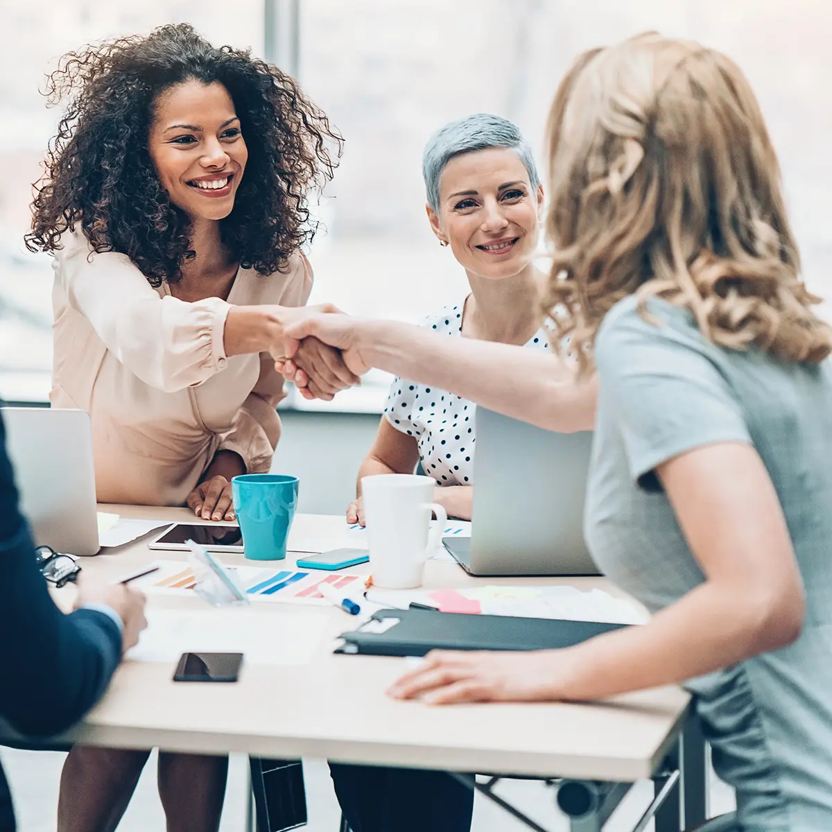Two smiling female colleagues shake hands across a table as another woman looks on.