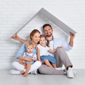 Family of four sits on floor against brick wall. Dad holds a roof cutout above them as kids sit on parents' laps.