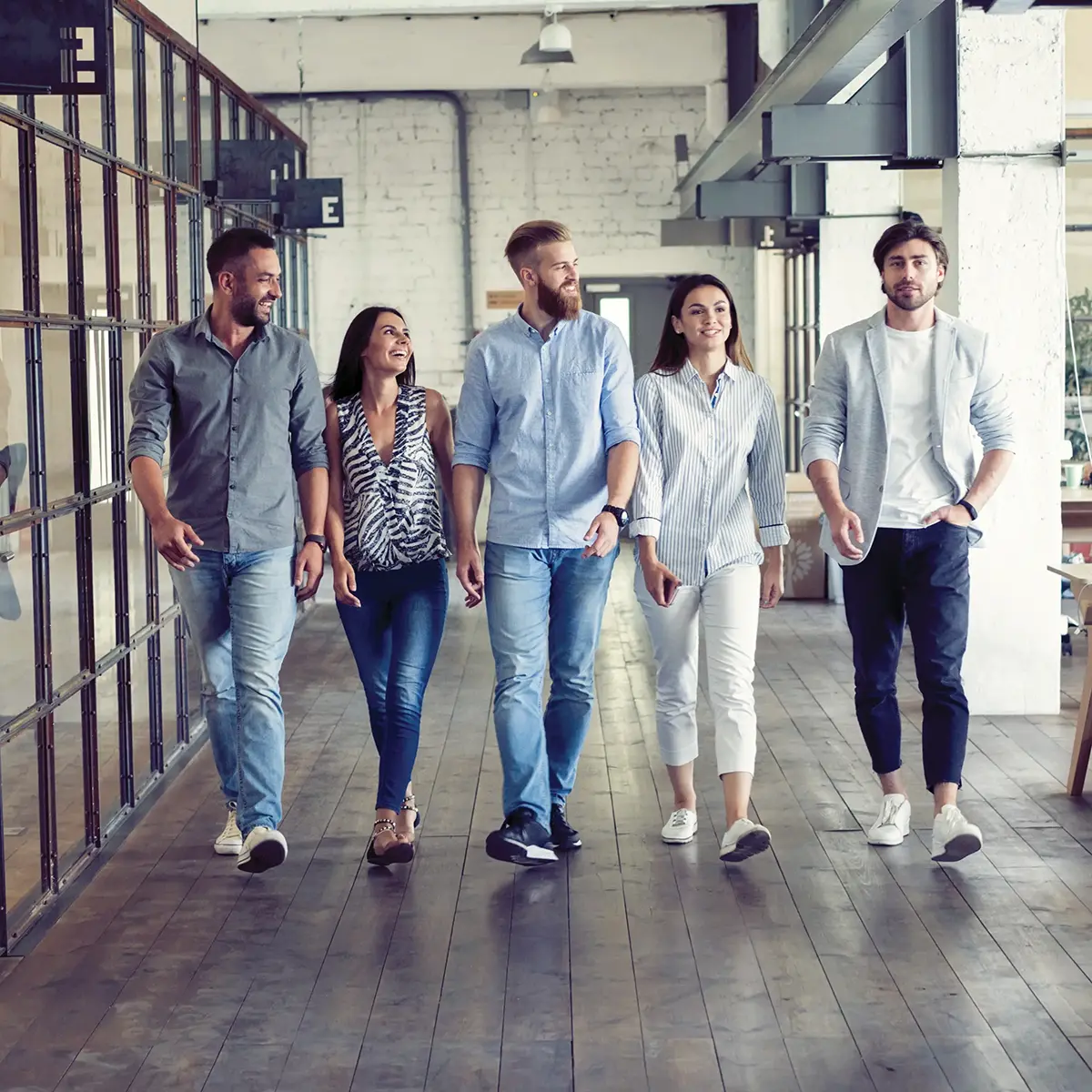 Five casually dressed individuals are walking and talking in a modern, industrial-style office space.
