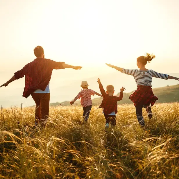Family of four—mom, dad, young daughter, and young son—run through a field of tall grass with arms outstretched
