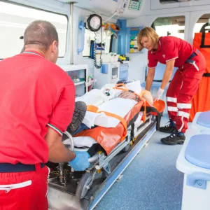 Man lies on a stretcher inside an ambulance, being assisted by a male and female EMT.