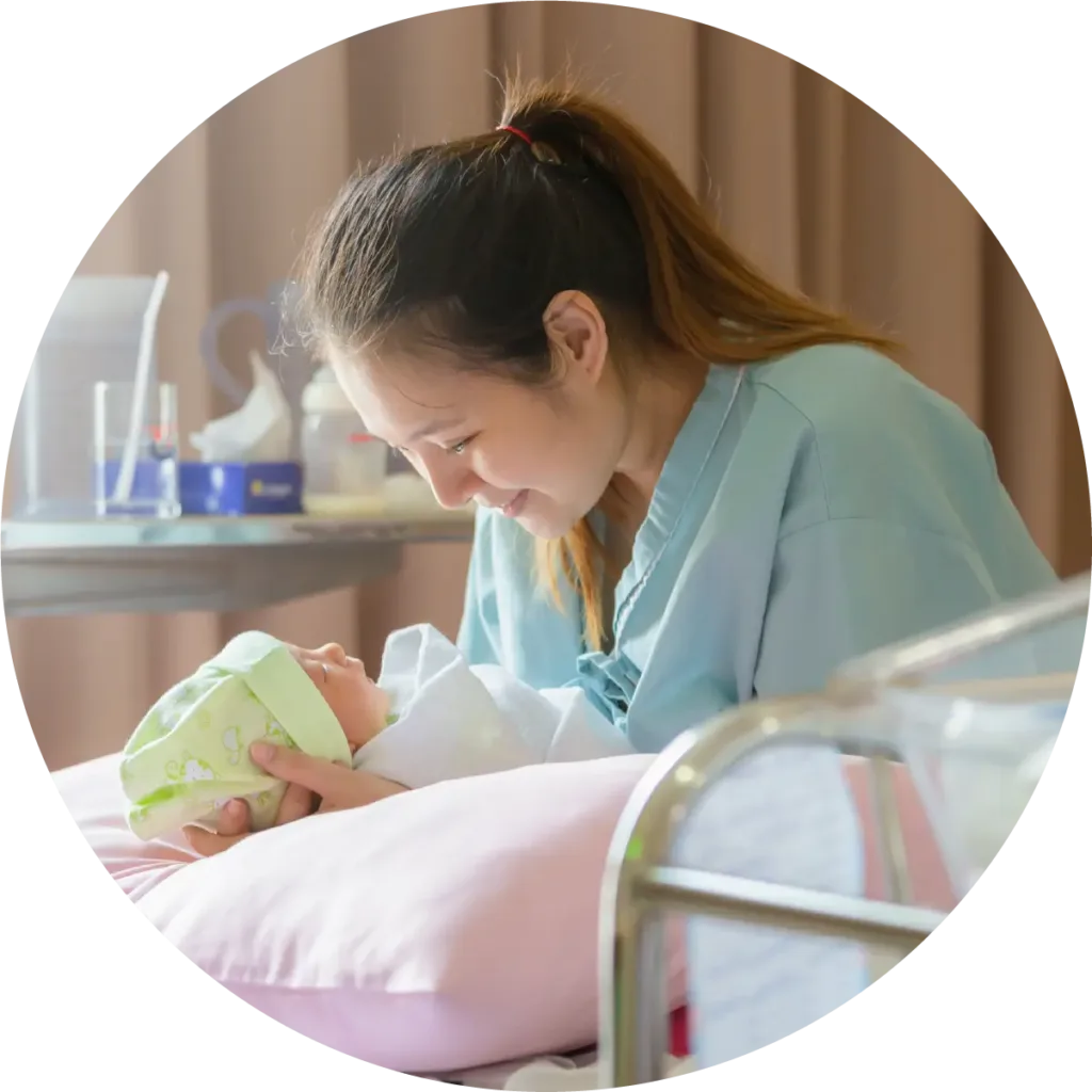 A mother lies in a hospital bed cradling her newborn baby’s head on a pillow, gazing at the baby with love.