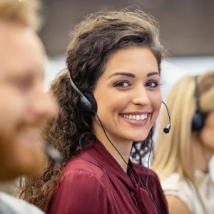 Smiling woman with a headset on, looking directly at the viewer in a call center environment.