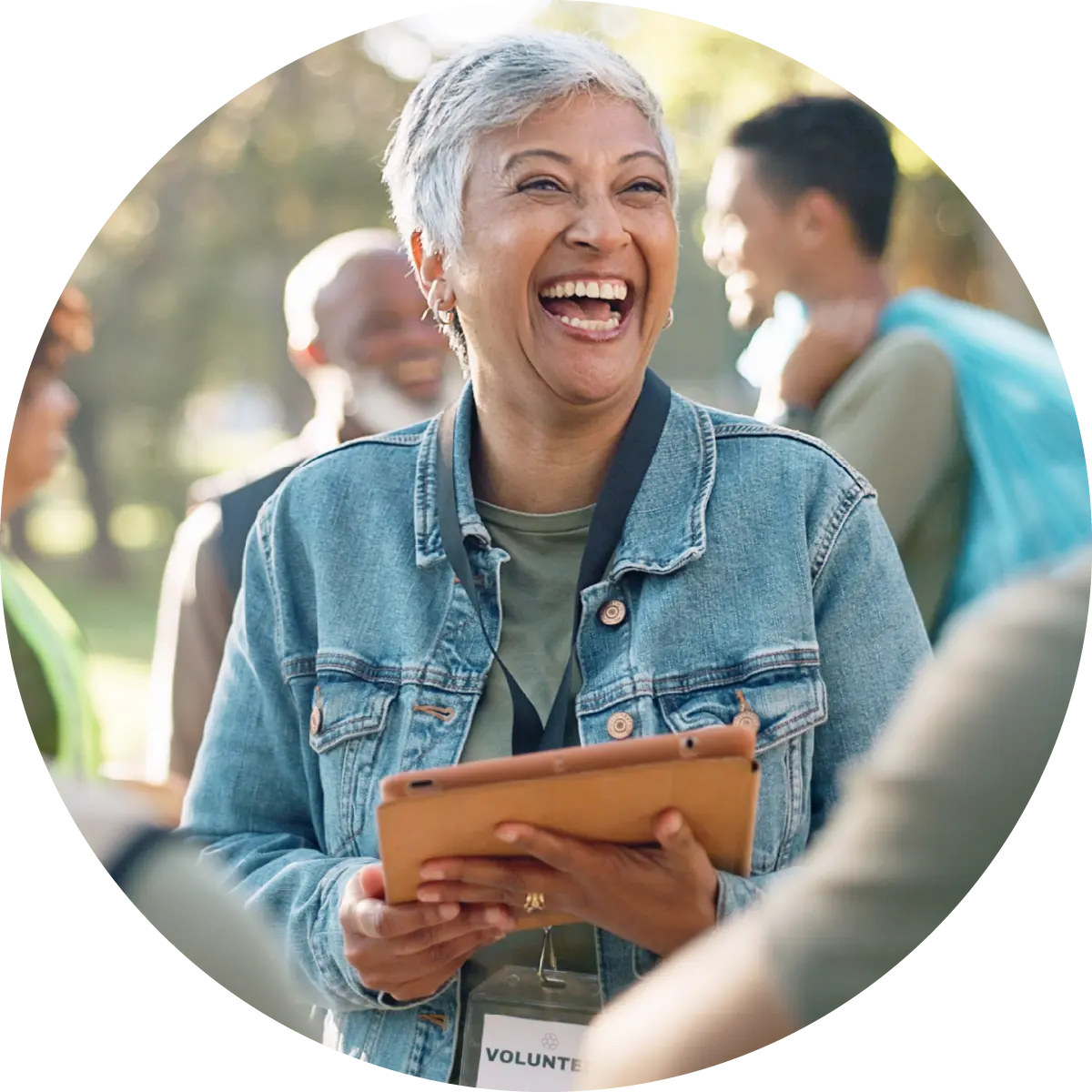 Joyful older female volunteer with short gray hair laughs while holding a tablet outdoors at an event.