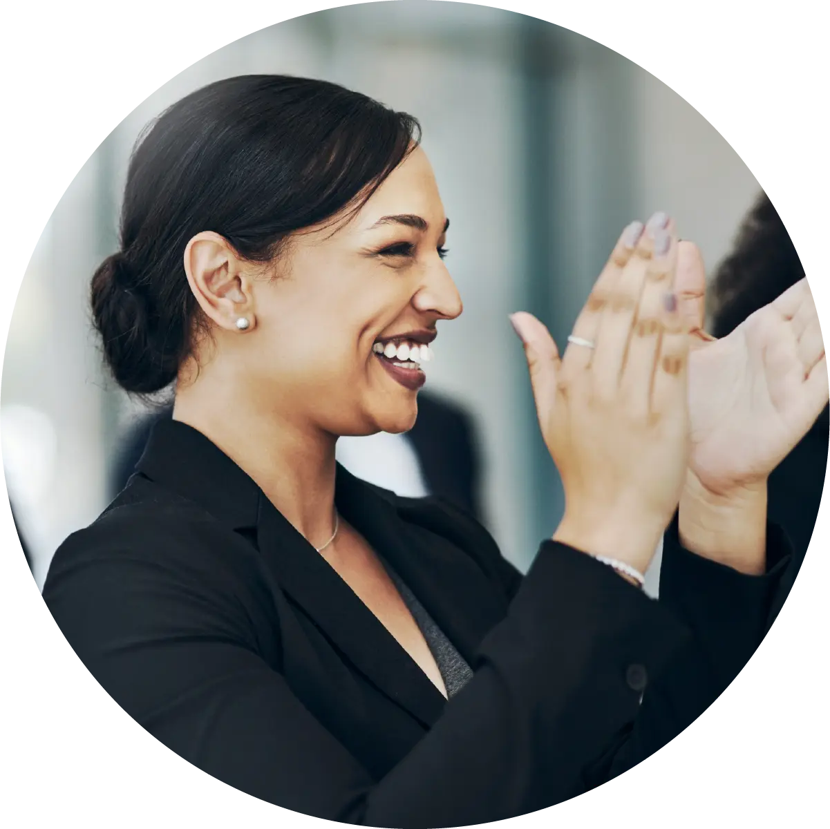 A smiling woman in a dark blazer shown in profile, clapping her hands with enthusiasm.