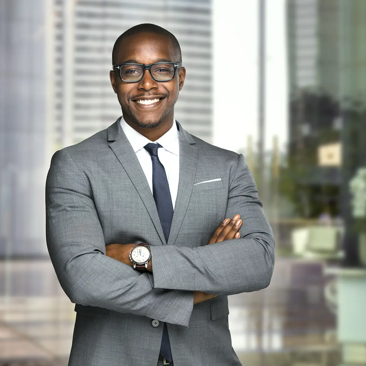 A smiling man with glasses, wearing a gray suit and tie, stands with his arms crossed in front of a blurred office background.