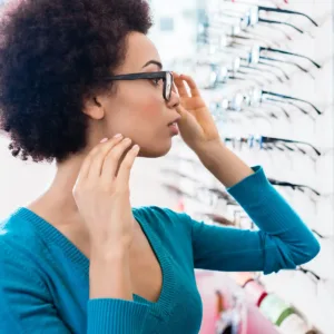 Woman stands by a wall of eyeglass frames in an optical store, trying on a pair of glasses in front of a mirror.