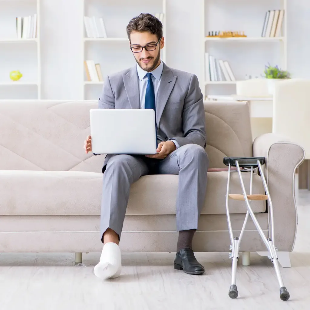 A man with a foot cast sits on a couch with crutches leaning nearby, looking at laptop resting on his lap.