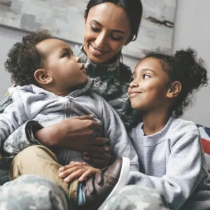 A woman in military uniform sits holding her son and daughter on her lap; both children smile up at her lovingly.